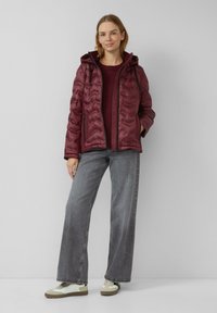 Femme se tenant contre un mur blanc, portant une veste matelassée bordeaux avec capuche, un pull bordeaux, un jean large gris et des baskets beiges.