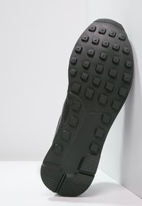 Suela de zapatilla negra con tacos de goma cuadrados y redondos, colocada en ángulo vertical contra una superficie blanca y fondo de pared.