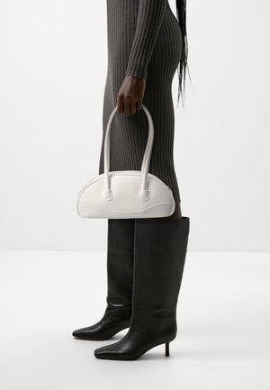 Kobieta w szarej prążkowanej sukience i czarnych kozaczkach do kolan trzyma białą, zaokrągloną torebkę za uchwyty na jednolitym tle.