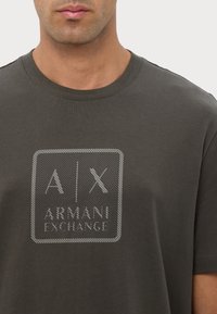 Camiseta de algodón gris oscuro con un logo cuadrado texturizado en blanco y gris, con el texto "ARMANI EXCHANGE" centrado debajo. Diseño de cuello redondo.