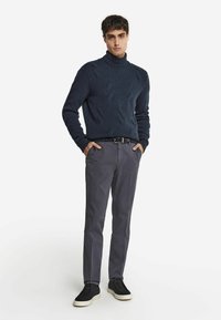 Marinblå stickad polotröja med texturerat mönster, kombinerad med grå byxor och svarta sneakers med krämfärgade sulor, stående hållning, händer i fickorna.