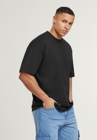 Black cotton T-shirt with a round neck and short sleeves, featuring a loose fit. Worn with light blue cargo shorts, hands in pockets.