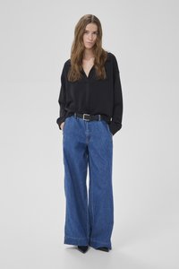 Donna con capelli lunghi castani che indossa una camicetta nera a maniche lunghe, jeans a gamba larga blu con cintura nera e scarpe nere con punta.