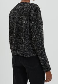 Pull noir texturé avec un motif tissé, col rond, manches longues et coupe légèrement courte. Vue arrière montrant l'ourlet et les poignets côtelés.