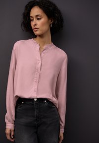 Blusa rosa chiaro a maniche lunghe con colletto arricciato e chiusura a bottoni, realizzata in un tessuto morbido, abbinata a jeans neri a vita alta.