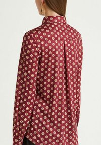Burgundy button-up shirt with a patterned design featuring small geometric shapes in beige and pink. Smooth fabric with a collar and cuffed sleeves.