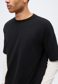 Νέος άνδρας με μαύρο κοντομάνικο μπλουζάκι πάνω από λευκό μακρυμάνικο πουκάμισο, στρέφεται προς τα αριστερά σε απλό φόντο.