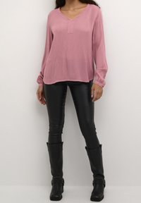 Blouse rose à manches longues avec un col en V et une texture plissée, associée à un pantalon moulant en simili cuir noir et des bottes noires montant jusqu'aux genoux.