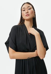 Musta kimono-tyylinen mekko, jossa on leveät hihat, valmistettu sileästä satiinimaisesta kankaasta. Mekossa on syvä V-pääntie ja virtaava siluetti, jossa on hienovarainen tekstuuri.