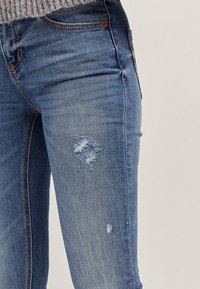 Blauwe spijkerbroek met een slanke pasvorm, met een verweerde textuur en rafelige plekken op het bovenbeen. Standaard vijfzakkenontwerp, medium wassing.