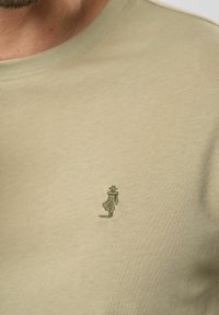 Khaki színű pamut póló kerek nyakkal és egy kisebb zöld hímzett figurával a bal mellrészen. Sima textúra, laza fazon.