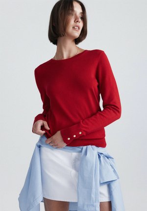 Femme portant un pull rouge à manches longues avec des boutons aux poignets, une jupe blanche et une chemise bleu clair nouée autour de la taille, regardant sur le côté.