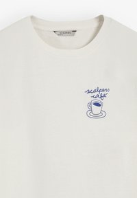 T-shirt beige clair avec col rond. Présente un graphisme bleu d'une tasse de café et le texte "scalpers café" sur la partie gauche de la poitrine.