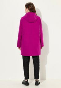 Fuchsia hættefrakke med sideslids, lige snit og blød tekstur, parret med sorte bukser og kraftige sorte sko.