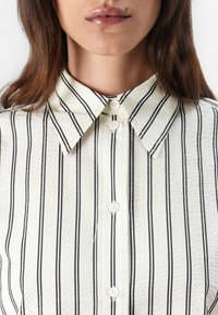 White shirt with black vertical stripes, textured fabric, classic collar, and button-up front detailing. Buttons are light-colored and round.