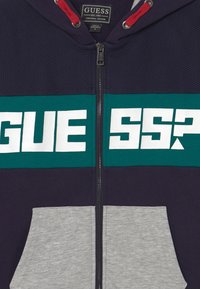 Sweat à zip bleu marine avec une bande verte, inscription "GUESS?" blanche, cordons rouges et poches avant grises.