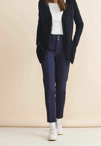 Marinblå byxor med skräddarsydd passform, försedd med elastisk midja och knappstängning, i kombination med en svart kofta och en vit topp.