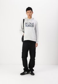 Grauer Hoodie mit blauer Grafikschrift, kombiniert mit schwarzen Cargo-Hosen mit Taschen. Schwarze Sneakers mit weißen Streifen. Minimalistischer Hintergrund.