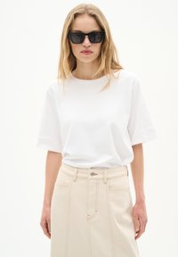 Weißes kurzärmliges Hemd kombiniert mit einem beigen, hoch-taillierten Rock, der über auffällige Nähte und einen Knopfverschluss verfügt. Schwarze Sonnenbrille runden das Outfit ab.