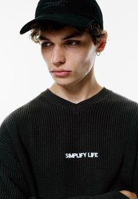 Pulover negru tricotat cu inscripția "SIMPLIFY LIFE" brodată în alb; design cu guler în V; modelul poartă o șapcă neagră; prim-plan care arată trăsăturile faciale și expresia.