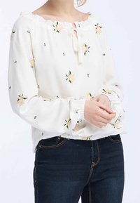 Blusa bianca a maniche lunghe con motivi floreali e piccoli ancore. Presenta una scollatura a chiave e dettagli con polsini elasticizzati.