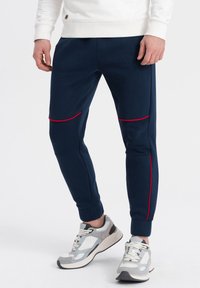 Marinblå joggingbyxor med texturerade detaljer och röda ränder, med avsmalnande ben och elastiska mudd. Grå sportskor synliga.