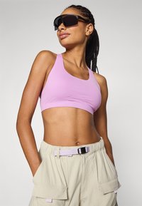Lys lilla sportbh laget av glatt stoff, med scoopet halslinning. Kombinert med beige cargo shorts, som har et teksturert belte.
