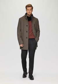 Bruine visgraatjas met zwarte knopen, gedragen over een bordeauxrood sweater. Slimme zwarte broek en zwarte leren laarzen maken de outfit compleet.