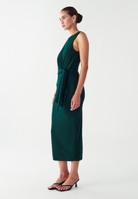 Žena u midi haljini bez rukava tamnozelene boje s remenom u struku, nosi zlatne naušnice, narukvice i crne sandale s visokom petom, stoji bočno.