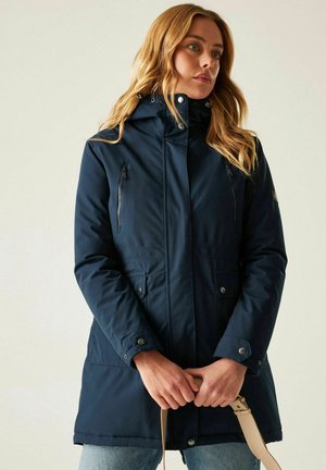 Mujer con parka azul marino con capucha, cremallera y bolsillos con botones, sosteniendo correas de bolso beige, de pie sobre un fondo claro y liso.