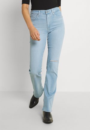Personne portant un jean taille haute bleu clair avec une petite déchirure au genou, un haut noir et des bottes noires à bout pointu, marchant sur un fond blanc uni.