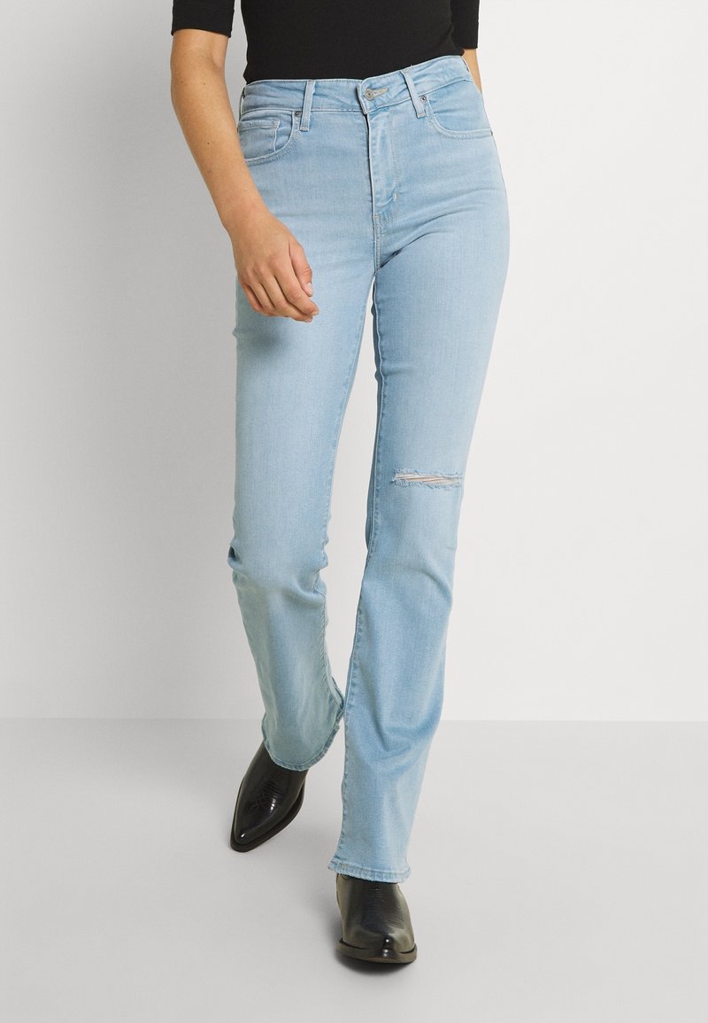 Personne portant un jean taille haute bleu clair avec une petite déchirure au genou, un haut noir et des bottes noires à bout pointu, marchant sur un fond blanc uni.