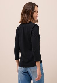 Haut noir à manches longues avec un col rond et une texture douce et lisse. Associé à un jean délavé, vu de dos.