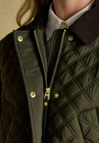 Zielona kurtka pikowana z ciemnym kołnierzem z kordurowej tkaniny, złotymi detalami metalowymi i zamkiem błyskawicznym, z widocznym wzorem w romby.