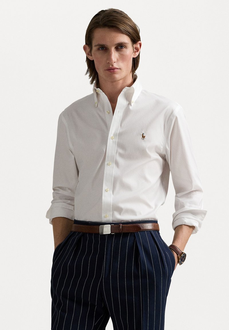 Chemise blanche à boutons avec un petit logo brodé, associée à un pantalon rayé bleu marine et une ceinture marron. Présente des manches retroussées.
