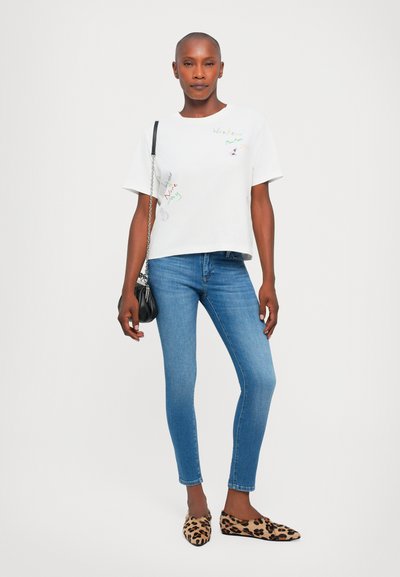 Vit kortärmad T-shirt med färgglad broderad text, tillsammans med blå skinnyjeans och leopardmönstrade ballerina-skor, hållande en svart handväska.