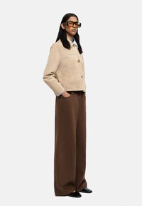 Beżowa zamszowa kurtka z dwoma guzikami i kieszeniami naszywanymi, w połączeniu z brązowymi spodniami szerokimi i czarnymi butami typu slip-on.