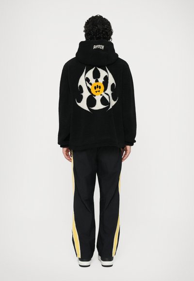 Veste polaire noire avec un grand motif blanc et jaune au dos. Associée à un pantalon noir avec des rayures latérales jaunes.