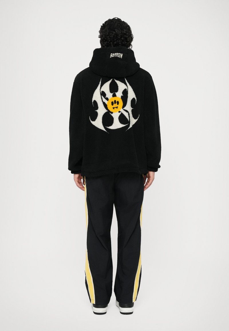 Fekete gyapjúdzseki nagy fehér és sárga grafikával a hátsó részen. Fekete nadrággal párosítva, melynek sárga oldalsávjai vannak.