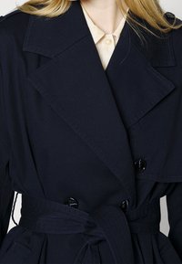 Námořnický trenčkot se zapínáním na dvouřadá knoflíky, velkými klopami, hladkou texturou a páskem z téhož materiálu. Černé knoflíky zdobí přední část.