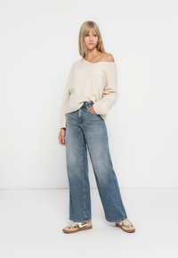 Hellebeige, schulterfreie Strickjacke kombiniert mit weit geschnittenen, verblichenen blauen Jeans und gemusterten beigen Turnschuhen. Minimalistischer weißer Hintergrund.