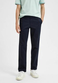 Navy blue chino pants in lightweight fabric, straight fit, featuring belt loops and side pockets, paired with white sneakers and light green shirt.