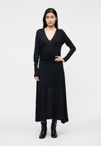 Μαύρο μακρυμάνικο φόρεμα με βαθύ V λαιμόκοψη, ρυτιδωμένες λεπτομέρειες στη μέση και ρέον φούστα. Κατασκευασμένο από ελαφρύ υλικό.