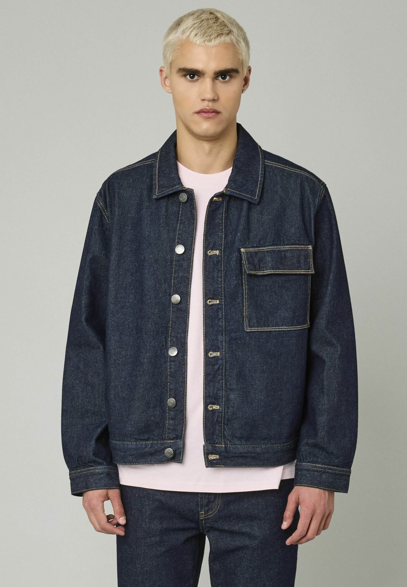 Veste en denim bleu foncé avec une fermeture à boutons, deux poches avant et des accents de couture jaunes. Associée à une chemise rose clair en dessous.