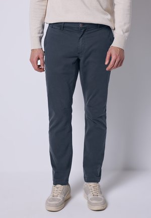 Pantaloni in cotone grigi con vestibilità slim, caratterizzati da una chiusura con bottone, tasche frontali e una trama sottile. Indossati con scarpe da ginnastica di colore chiaro.