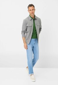 Camicia grigia con bottoni e due tasche sul petto, indossata sopra un polo verde, abbinata a jeans azzurri chiari e sneakers beige. Design semplice e contemporaneo.
