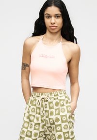 Γυναίκα με μακριά μαύρα μαλλιά, που φοράει ένα ανοιχτό ροζ κορυφαίο τοπ τύπου halter με την επιγραφή «Santa Cruz» και πράσινα σορτς με σχέδια, με τα χέρια στις τσέπες.