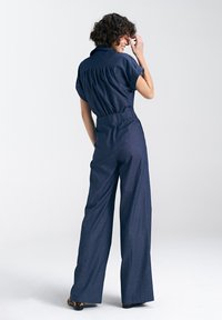 Granatowy dżinsowy kombinezon z krótkimi rękawami, paskiem w talii i szerokimi spodniami. Posiada kołnierzyk oraz marszczenia z tyłu dla luźniejszego kroju.