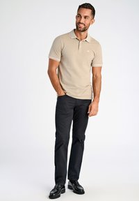 Camisa polo beige con ribete blanco, con un pequeño logo en el pecho. Combinada con pantalones negros y zapatos negros. Diseño sencillo, de mangas cortas.