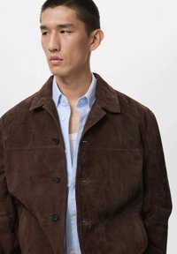 Giacca in suede marrone con colletto classico, dotata di cinque bottoni neri e due tasche laterali, indossata sopra una camicia azzurra chiara.